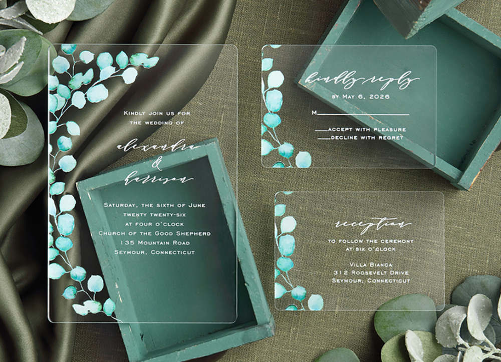 A clear acrylic wedding invitation with a green watercolor eucalyptus design and white ink wording