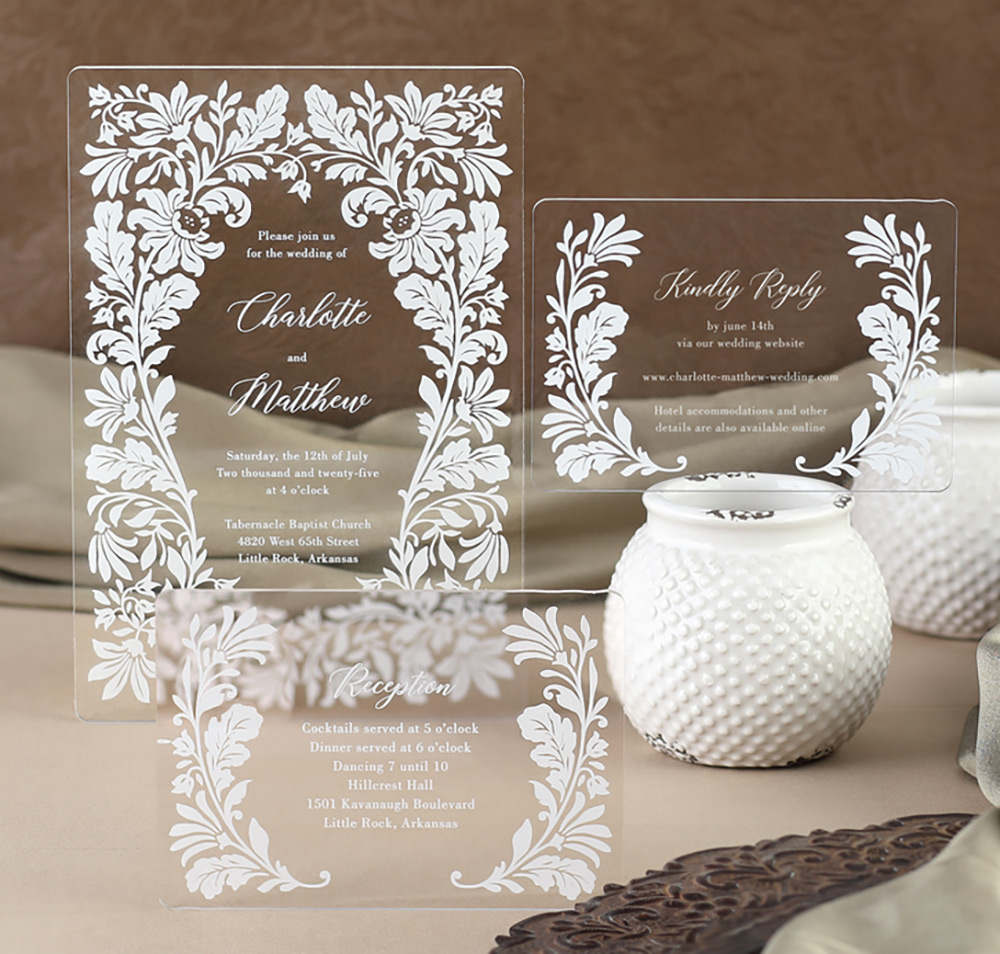 A floral design printed in white ink frames the wording on a clear acrylic wedding invitation