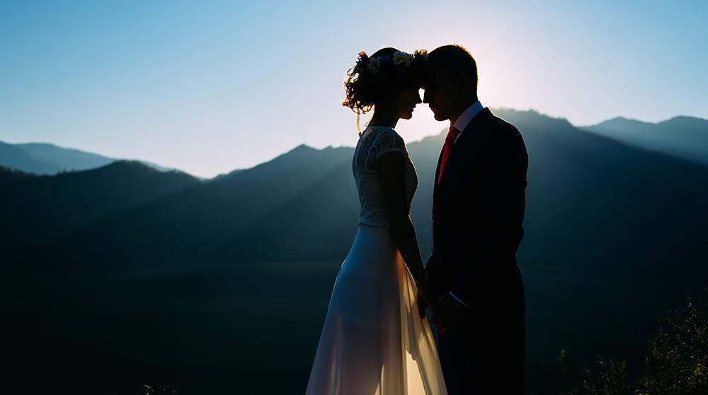 A bride and groom stand silhouetted against a mountain range