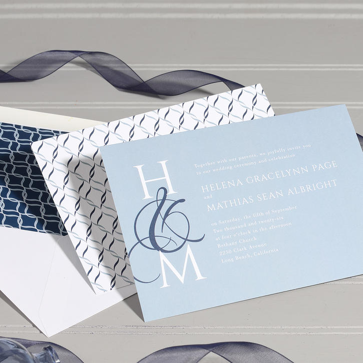 A light blue wedding invitation featuring a modern monogram with a dark blue ampersand shown with coordinating envelopes