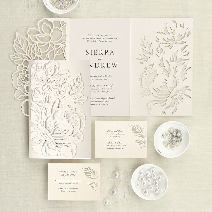 An ecru shimmer wrap laser cut with an intricate floral design wraps around a matching wedding invitation and is shown with two matching enclosures