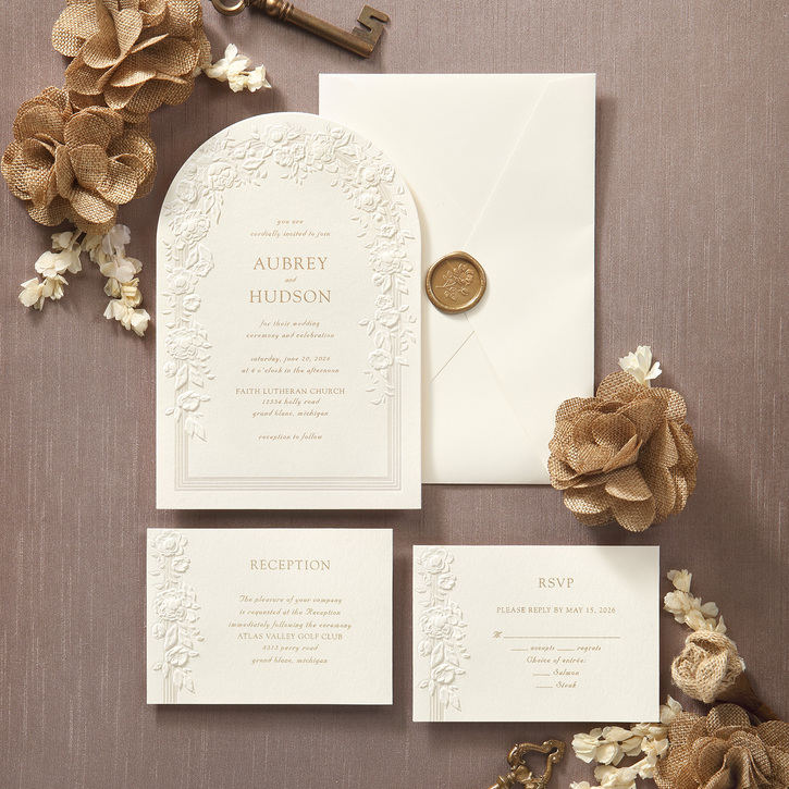 A classic wedding invitation in ecru with floral embossing shown with matching ensemble pieces such as enclosures, envelope and a wax seal