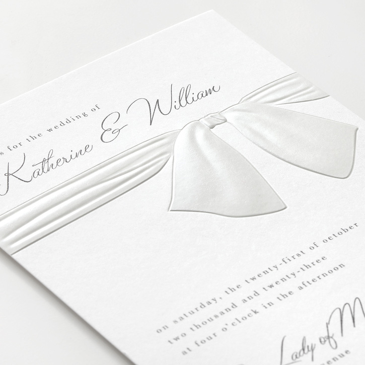 A wedding invitation featuring pearl embossed bow at the top. 